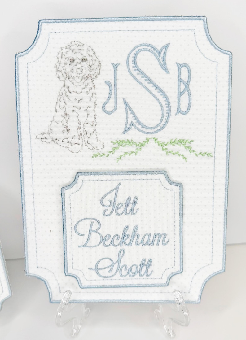 May include: Embroidered white fabric plaque with a light blue border and polka dot pattern. Features a dog illustration, initials, and the name "Jett Beckham Scott" in script. Includes a clear plastic stand.