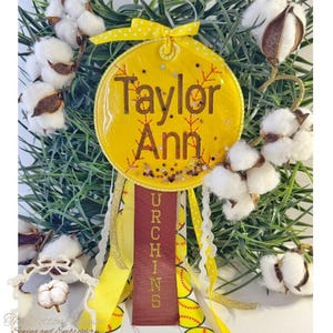 May include: A yellow softball-themed ornament with the name "Taylor Ann" in brown lettering. The ornament has a ribbon with the word "URCHINS" and is surrounded by cotton and greenery.