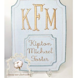 May include: A personalized, light blue and white gingham-patterned plaque with gold embroidered initials "KFM". Below, embroidered text reads "Kipton Michael Foster". The plaque has a decorative border and is from "The Cotton House Sewing and Embroidery".