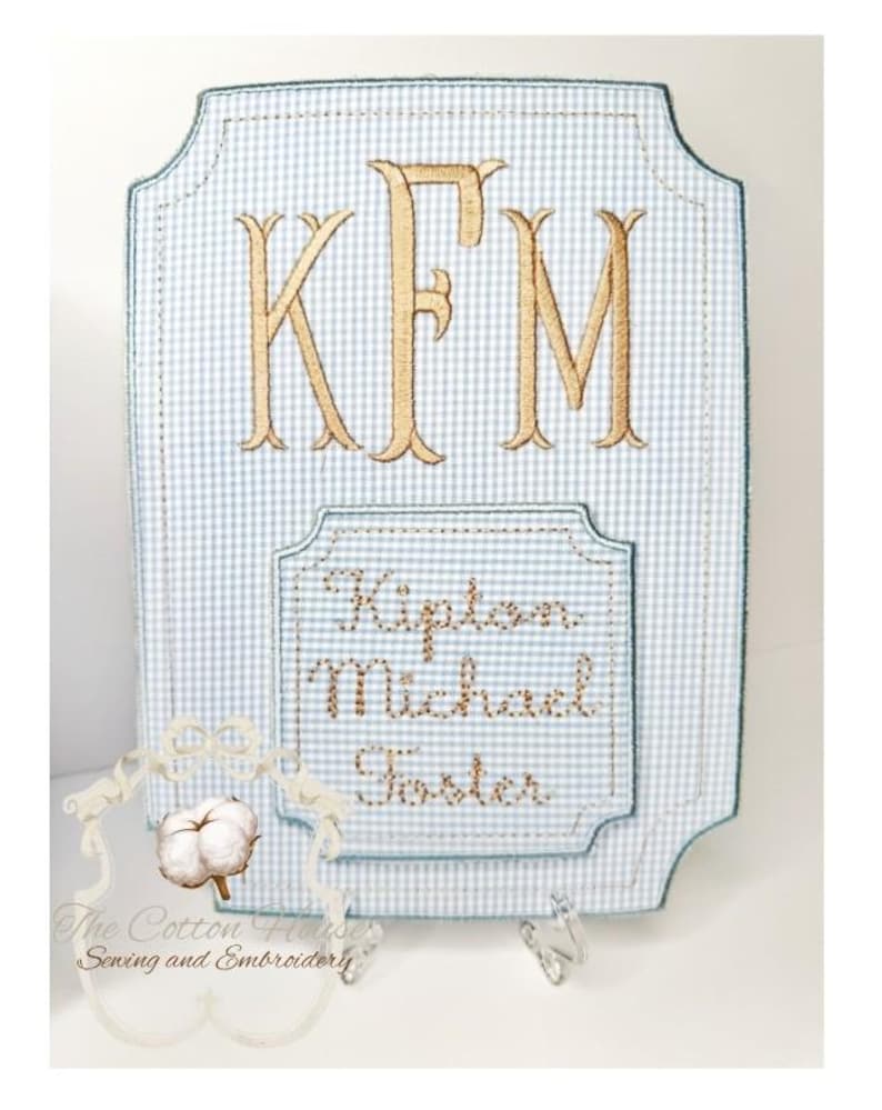 May include: A personalized, light blue and white gingham-patterned plaque with gold embroidered initials "KFM". Below, embroidered text reads "Kipton Michael Foster". The plaque has a decorative border and is from "The Cotton House Sewing and Embroidery".