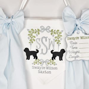 May include: A white banner with a light blue bow on each side. The banner has a floral wreath design with the initials "SSW" and two black dog silhouettes. A tag with the name "Swayze Wilson" is attached.