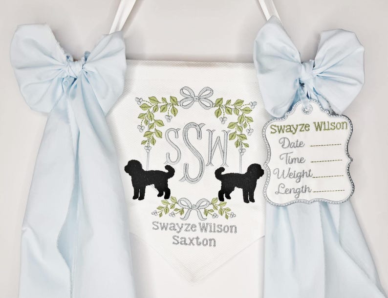 May include: A white banner with a light blue bow on each side. The banner has a floral wreath design with the initials "SSW" and two black dog silhouettes. A tag with the name "Swayze Wilson" is attached.
