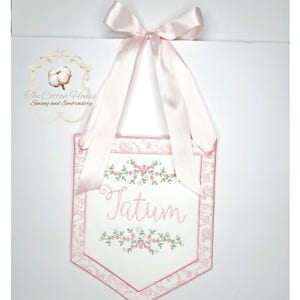 May include: A white and pink embroidered banner with the name "Tatum" in cursive script. The banner features floral embroidery and a pink ribbon for hanging. The Cotton House Sewing and Embroidery logo is visible.