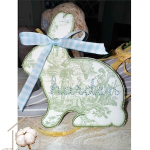 May include: A decorative Easter bunny-shaped ornament. The ornament features a green and white toile pattern with a blue and white gingham ribbon bow. The edges are outlined in green. The Cotton House logo is in the bottom left corner.