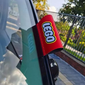 Könnte beinhalten: Ein rotes und gelbes LEGO Banner ist an einer Autoscheibe befestigt. Das LEGO-Logo ist in Weiß und Gelb auf dem roten Banner deutlich sichtbar. Das Auto ist schwarz, und der Hintergrund zeigt Bäume und einen blauen Himmel.