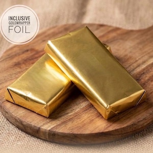 May include: Two rectangular bars wrapped in shiny gold foil. A circular graphic in the upper left corner contains the text INCLUSIVE GOLDWRAPPER FOIL. The bars are positioned on a wooden surface with a visible grain and a rounded handle.