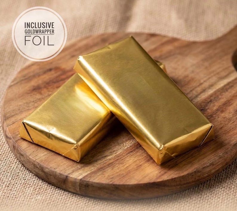 May include: Two rectangular bars wrapped in shiny gold foil. A circular graphic in the upper left corner contains the text INCLUSIVE GOLDWRAPPER FOIL. The bars are positioned on a wooden surface with a visible grain and a rounded handle.