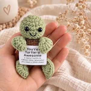 Könnte beinhalten: Eine kleine gehäkelte Schildkröte aus salbeigrünem und braunem Garn mit schwarzen Kunststoffaugen. Die Schildkröte hält eine weiße rechteckige Karte mit dem aufgedruckten Text: You're Turtle-y Awesome. A teeny turtle is always here, whenever you need a little cheer.