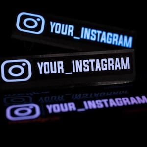 Könnte beinhalten: Zwei rechteckige schwarze Aufkleber mit dem weißen Instagram-Kamerasymbol auf der linken Seite und dem weißen Text YOUR_INSTAGRAM auf der rechten Seite. Die Aufkleber sind vor einem dunklen Hintergrund mit einer Spiegelung darunter platziert.