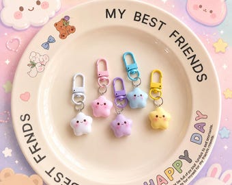 May include: Five star-shaped keychains in white, pink, purple, blue, and yellow, each featuring a smiling face with rosy cheeks. Each keychain has a matching colored lobster clasp attached by a silver metal ring, resting on a cream-colored plate.