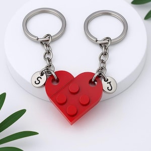 Könnte beinhalten: Zwei rote herzförmige Schlüsselanhänger. Jeder Schlüsselanhänger hat einen silbernen Ring, eine Kette und einen Anhänger mit einem Buchstaben. Ein Anhänger hat den Buchstaben "S" und der andere den Buchstaben "J". Das Herz besteht aus zwei ineinandergreifenden Teilen.