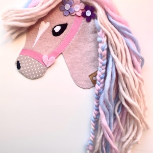 Könnte beinhalten: Ein Pferdekopf aus Filz in zartem Rosa mit einem rosa Halfter und einer gepunkteten Schnauze. Die Mähne ist eine Mischung aus rosa, blauem und weißem Garn, mit einem geflochtenen Abschnitt. Der Pferdekopf ist mit Filzblumen verziert.