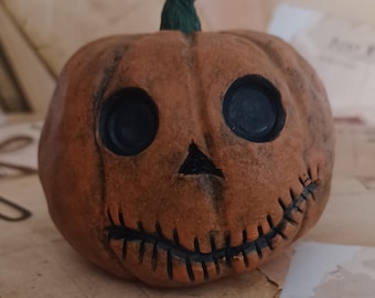 Può includere: Una piccola zucca arancione con un sorriso cucito e grandi occhi neri. La zucca ha uno stelo verde e un aspetto usurato e vintage.