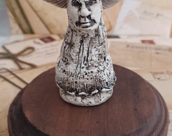 Puede incluir: Una figurita de cerámica de un hongo con cara humana. El hongo está pintado de blanco con detalles marrones y tiene un sombrero rojo con manchas blancas. La figurita está sentada sobre una base de madera.