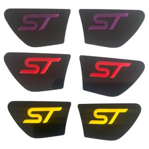 May include: Six black car emblems featuring the letters "ST" in purple, red, and yellow. The emblems have a glossy finish and a curved, trapezoid shape. The "ST" letters are raised and have a metallic appearance.