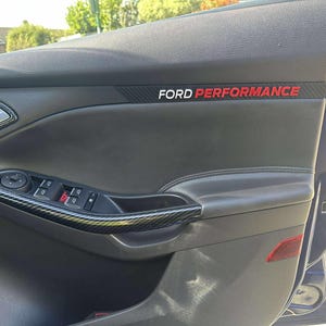 May include: Dark grey car door panel with carbon fibre trim. The words "FORD PERFORMANCE" are printed in white and red. The door panel features window and mirror controls.