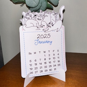 May include: A white desk calendar with a stand featuring a group of cats in a pile. The calendar is for January 2025 and has the days of the week labeled at the top.