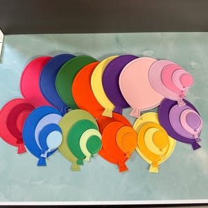 May include: A collection of colorful, layered paper balloon cutouts in various sizes and shades. The balloons are in shades of red, blue, green, orange, yellow, and purple. Each balloon has a stitched outline and a small, attached string.