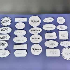 May include: A collection of white paper cutouts with black text on a purple surface. The cutouts have various shapes, including circles, ovals, and rectangles. The text includes phrases like "Praying for you" and "You've been in my prayers."