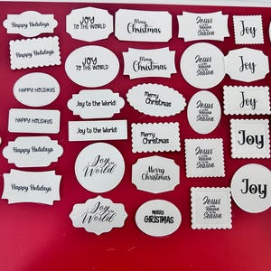 May include: A collection of white paper cutouts with various holiday phrases in black script, including "Seasons Greetings," "Happy Holidays," "Joy to the World," and "Merry Christmas." The cutouts are in different shapes, such as rectangles, ovals, and circles, and are arranged on a red surface.