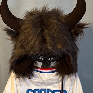 Può includere: Un cappello di bufalo in finta pelliccia marrone con grandi corna marroni e paraorecchie. Il cappello è su una maglia bianca con il nome "COOPER" in lettere blu.
