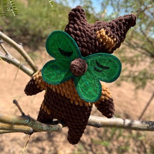 May include: A handmade crochet doll in brown and tan with a green felt clover. The doll has a whimsical design, with a clover attached to its chest. The doll is perched on a tree branch, with a blurred natural background.