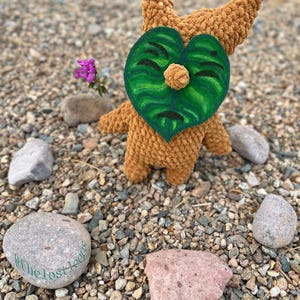 May include: A handmade brown crochet doll with a green leaf-shaped face and a small, round nose. The doll has two small horns and is surrounded by small rocks and pebbles. A small purple flower is visible in the background.