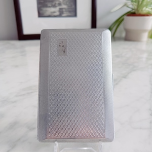 May include: A silver rectangular cigarette case with a textured, diamond-patterned surface. The case features the initials "A S" in a square on the front. The case is displayed on a clear stand.