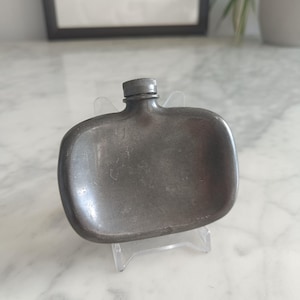 May include: A small, rectangular, metallic flask with rounded corners and a smooth, dark grey finish. The flask features a centred, short, cylindrical neck topped with a small, ribbed, screw-on cap, resting on a clear plastic display stand.