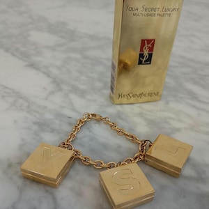 May include: A gold-toned charm bracelet with three square charms spelling "YSL". A gold box with the text "Your Secret Luxury Multi-Usage Palette" and the Yves Saint Laurent logo is also in the image.