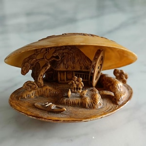 Könnte beinhalten: Eine beigefarbene, geschnitzte Muschelskulptur mit einer detaillierten Miniaturlandschaft im Inneren. Die Szene zeigt ein kleines Haus mit Strohdach, ein Wasserrad, zwei Figuren, ein Boot und Bäume, die kunstvoll in das Innere eingearbeitet sind.