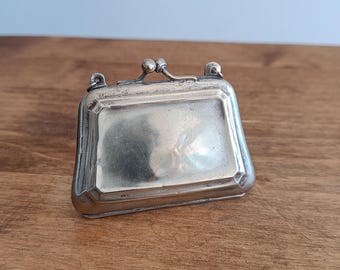 Puede incluir: Un pequeño bolso de plata con un cierre de bisagra y un asa decorativa. El bolso está cerrado y se encuentra sobre una superficie de madera.