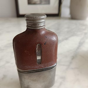 Könnte beinhalten: Ein Flachmann mit einem braunen, lederummantelten Oberteil, das ein vertikales Sichtfenster aus Glas aufweist. Der untere Teil besteht aus gebürstetem, silberfarbenem Metall und ist mit einem passenden Schraubverschluss mit Rillen versehen.