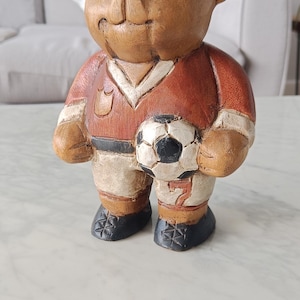 May include: A wooden bear figurine wearing a red and white soccer uniform, holding a black and white soccer ball. The bear has a carved appearance with visible wood grain and is standing on a white surface.