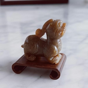May include: A detailed, amber-colored jade figurine of a mythical creature, possibly a Pixiu, on a dark wooden stand. The sculpture has intricate carvings and sits on a curved, rectangular base. The background is a blurred, neutral tone.
