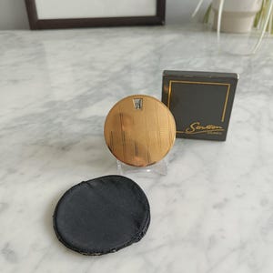 May include: A vintage gold-toned Stratton compact with a textured design and the letter "A" on the lid. A black powder puff and a black box with gold trim and the name "Stratton London" are also present.