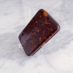 Puede incluir: Un estuche rectangular con un patrón de carey y un acabado brillante. El estuche es de color marrón oscuro con veteado marrón claro y naranja. Se encuentra sobre un soporte de exhibición transparente inclinado.