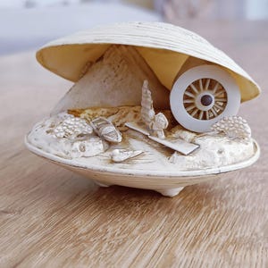 Könnte beinhalten: Ein cremefarbenes, muschelförmiges Dekorationsobjekt mit einer Miniaturlandschaft im Inneren. Die Landschaft umfasst ein Boot, Bäume und ein Rad. Das Objekt steht auf einer Holzoberfläche.