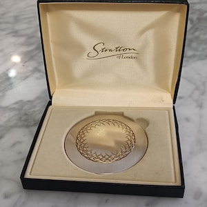 May include: A vintage Stratton of London compact mirror in a black box. The compact has a gold-toned, patterned design on a silver-coloured base. The box is lined with a cream-coloured fabric and has the Stratton of London logo in gold.