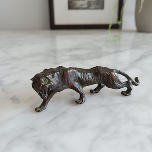 Op de afbeelding: Een klein metalen beeldje in een donkere bronskleur van een sluipende tijger in een lage, agressieve houding. Het beeldhouwwerk heeft een gedetailleerde vachtstructuur, een grommende bek met zichtbare tanden en een lange, gebogen staart.