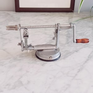 May include: A vintage silver apple peeler with a wooden handle. The kitchen tool has a suction base and a crank for peeling apples. A framed black and white photo hangs in the background.