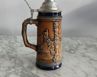May include: A brown and blue ceramic beer stein with a silver lid. The stein features a relief design of a man on horseback and the text "ANDREAS HOFER".