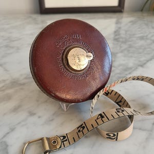 Puede incluir: Un metro de cinta de cuero marrón vintage con el texto "CONSTANTIA SHEFFIELD ENGLAND" y un mecanismo de bobinado de metal. La cinta beige está marcada con números negros y se extiende hasta 1,52 metros.