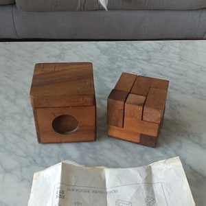 Pode incluir: Dois quebra-cabeças de cubo de madeira, um montado e outro desmontado, estão em uma superfície de mármore. Um diagrama com instruções está abaixo. Um cão preto e branco descansa em um sofá cinza ao fundo.