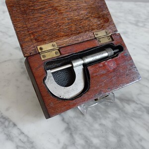 May include: A silver micrometer in a wooden box with brass hinges. The micrometer has a black leather covering on the handle. The box is open and the micrometer is inside.