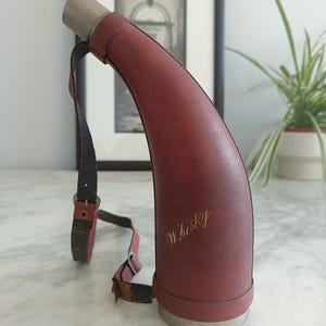 May include: A curved, horn-shaped flask covered in reddish-brown leather with a metallic base and cap. The word "Whisky" is embossed in cursive script on the side. A worn, adjustable leather strap with a metal buckle is attached to the flask.