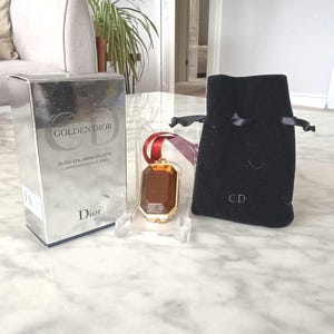 May include: A silver box with "GOLDEN DIOR" text and a makeup jewel. The jewel is a rectangular shape with a gold frame and a red ribbon. A black velvet pouch with "CD" is also present.