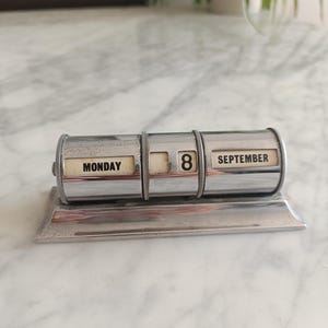 Puede incluir: Un calendario de escritorio vintage de cromo con tres secciones cilíndricas giratorias. La pantalla muestra el texto MONDAY, el número 8 y el texto SEPTEMBER en paneles rectangulares de color blanco roto sobre una base de marco metálico pulido.