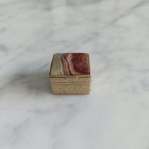 May include: A small square metal box with a hinged lid featuring a polished stone inlay with swirling bands of red, white, and gray. The gold-toned metal sides are decorated with an embossed floral pattern and a twisted rope border.