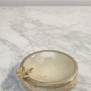 Puede incluir: Un peque&ntilde;o cenicero redondo de alabastro blanco con un dise&ntilde;o de hoja de oro. El cenicero es redondo y tiene un borde ligeramente elevado.